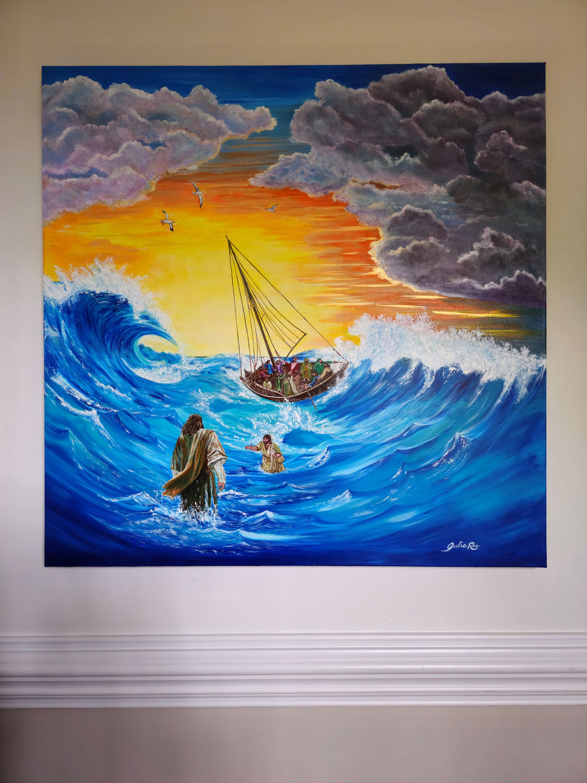 The Storm artwork by Julie Rae displayed on a beige wall, showing Jesus and Peter amid crashing blue waves.
